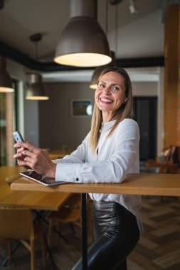 Yetişkin bir orta yaşlı beyaz kadın iş yerinde mutlu bir gülümseme takınıyor, beyaz gömlek giyiyor, ön plan fotoğrafını cep telefonuyla çekiyor ya da kendine güvenen mesajlar yolluyor.