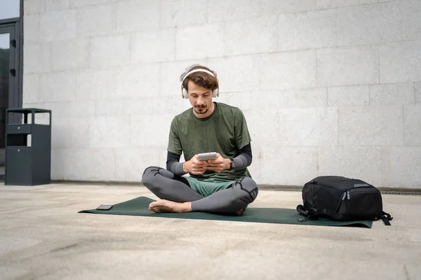 stock image One man young adult caucasian male prepare for guided training yoga or meditation hold digital tablet while sitting outdoor with headphones self-care practice well-being inner peace balance concept