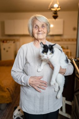 Yaşlı, yaşlı, yaşlı, beyaz, yaşlı bir kadın, evde tek başına duruyor. Belden yukarı portre, kameraya bak. Kedi evcil hayvanı, bunama hastalığı ve depresyon konsepti.