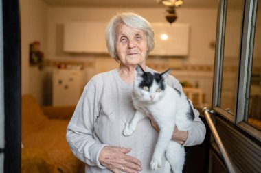 Yaşlı, yaşlı, yaşlı, beyaz, yaşlı bir kadın, evde tek başına duruyor. Belden yukarı portre, kameraya bak. Kedi evcil hayvanı, bunama hastalığı ve depresyon konsepti.