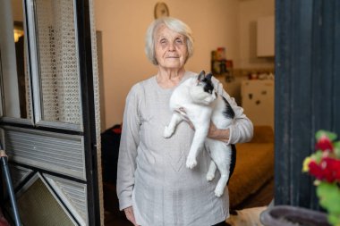 Yaşlı, yaşlı, yaşlı, beyaz, yaşlı bir kadın, evde tek başına duruyor. Belden yukarı portre, kameraya bak. Kedi evcil hayvanı, bunama hastalığı ve depresyon konsepti.