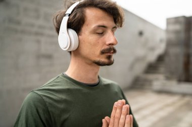 Genç, yetişkin, beyaz, bıyıklı bir erkek portresi. Kulaklık takıyor, kameraya bakıyor. Gerçek insanlar uzay belini açık havada meditasyon konseptini taklit ediyor.