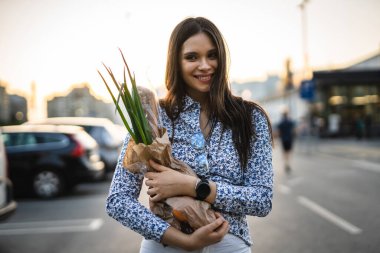 Bir kadın, alışveriş merkezinin önündeki otoparkta yürürken ya da ayakta dikilirken elinde yiyecek taşıyan genç ve güzel bir esmerin portresini yukarı kaldırıyor.