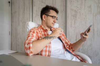 Kafkasyalı bir erkek, çalışma masasındaki ofiste otururken cep telefonuyla arama yapıyor. Proje üzerinde çalışırken gömlek ve gözlük takıyor.