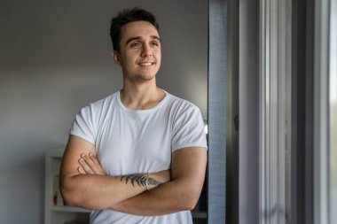 One man young adult male caucasian with brown hair standing by the window at home in day confident looking to the side happy smile wear white shirt real people copy space