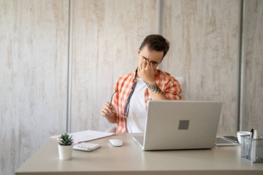 One man sitting at the desk at work in the office in chair tired overworked and exhausted having eyestrain due long usage of laptop computer hold eyeglasses real people freelancer eye pain hold head