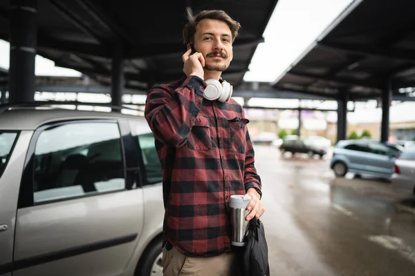 携帯電話のスマートフォンの話を使用して一日の駐車場に立っている一人の若い大人の白人男性一人が一日で一人で待つ仕事に行きながら 実際の人々はスペースをコピーしますホールドブリーフケースとコーヒーカップ — ストック写真
