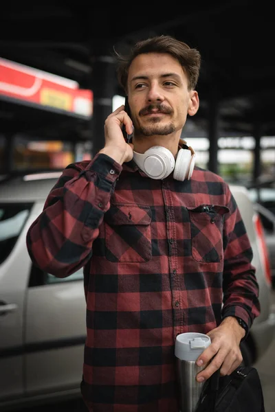 携帯電話のスマートフォンの話を使用して一日の駐車場に立っている一人の若い大人の白人男性一人が一日で一人で待つ仕事に行きながら 実際の人々はスペースをコピーしますホールドブリーフケースとコーヒーカップ — ストック写真