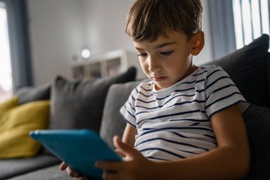 Kafkasyalı bir anaokulu öğrencisi dijital tablette oyun oynuyor. Çocukluk çağında, modern yaşam tarzı konseptinin fotokopisini çekerken çocuk gelişimi kanepede oturuyor.