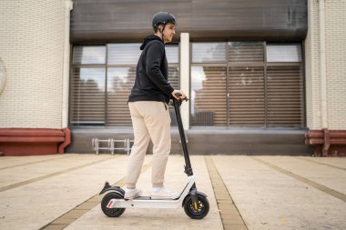 Genç, yetişkin, beyaz bir erkek. Elektrikli scooterın üzerinde duruyor. Şehirde ya da şehirde araba sürüyor.