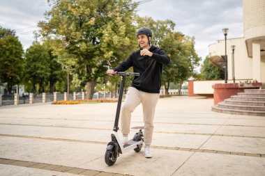 Genç, yetişkin, beyaz bir erkek. Elektrikli scooterın üzerinde duruyor. Şehirde ya da şehirde araba sürüyor.
