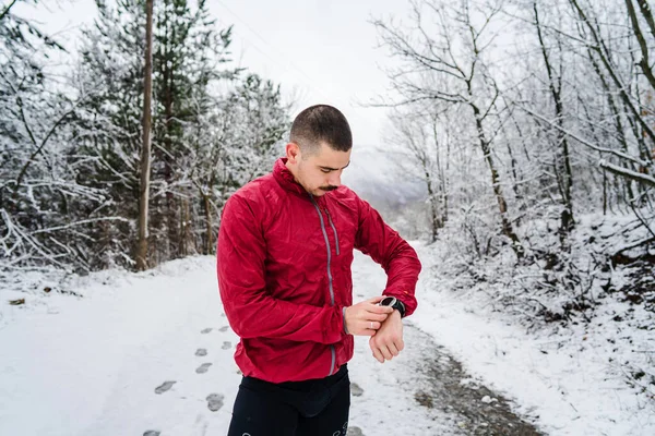 One adult man standing outdoor in winter day checking his smart wristwatch for time and hart-rate while training outdoor male athlete runner using mobile app while training in winter day in nature
