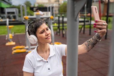 Bir kadın olgun ya da yaşlı beyaz kadın eğitimi. Açık spor salonunun parkındaki basın fitness makinesi üzerinde cep telefonu uygulaması. Sağlıklı yaşam tarzı konsepti.