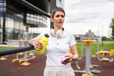 Beyaz bir kadın açık hava antrenmanı sırasında fren yapıyor. Açık hava spor salonunda barlarda dinleniyor. İçme suyu ya da takviye malzemesi ile birlikte.