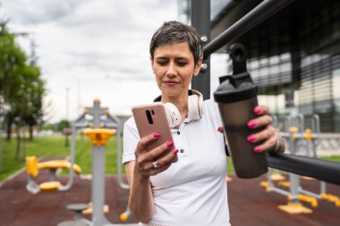 Beyaz bir kadın açık hava antrenmanı sırasında fren yapıyor. Açık hava spor salonunda barlarda dinleniyor. İçme suyu ya da takviye su ile dinleniyor.