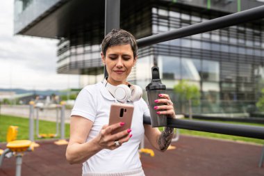 Beyaz bir kadın açık hava antrenmanı sırasında fren yapıyor. Açık hava spor salonunda barlarda dinleniyor. İçme suyu ya da takviye su ile dinleniyor.