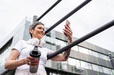 Beyaz bir kadın açık hava antrenmanı sırasında fren yapıyor. Açık hava spor salonunda barlarda dinleniyor. İçme suyu ya da takviye su ile dinleniyor.