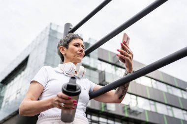 Beyaz bir kadın açık hava antrenmanı sırasında fren yapıyor. Açık hava spor salonunda barlarda dinleniyor. İçme suyu ya da takviye su ile dinleniyor.