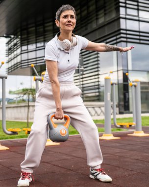 Bir kadın, modern olgun, beyaz kadın. Kısa saçlı, bina önünde günün ilerleyen saatlerinde spor salonunda kettlebell ağırlığı ile spor salonu sağlıklı yaşam tarzı konseptini taklit ediyor.