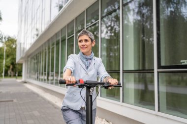 Yetişkin bir son sınıf beyaz kadın elektrik tekmeli scooter 'a biniyor. Şehirde ya da şehirde araba sürüyor. E-scooter çevre dostu.