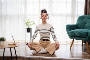Yetişkin bir beyaz kadın milenyum kulaklıklarını internet rehberli meditasyon için kullanıyor. Evde gözleri kapalı farkındalık yogası yapıyor. Gerçek insanlar kişisel bakım kavramını taklit ediyor.