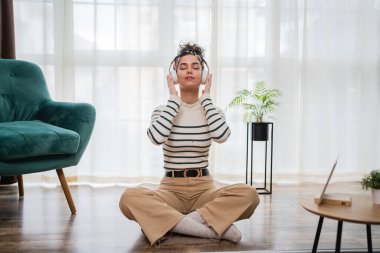 Yetişkin bir beyaz kadın milenyum kulaklıklarını internet rehberli meditasyon için kullanıyor. Evde gözleri kapalı farkındalık yogası yapıyor. Gerçek insanlar kişisel bakım kavramını taklit ediyor.