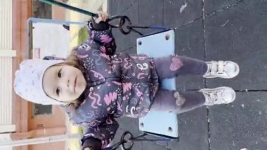 Bir kız çocuğu, iki yaşında, kış ya da sonbahar günü şehirde salıncakta sallanıyor. UGC kullanıcısı, içerik olarak dikey otantik video üretti.