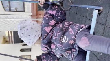 Bir kız çocuğu, iki yaşında, kış ya da sonbahar günü şehirde salıncakta sallanıyor. UGC kullanıcısı, içerik olarak dikey otantik video üretti.