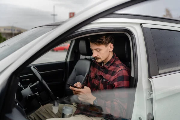 One man caucasian male with mustaches sit in the car take a brake on road-trip hold cup of coffee use mobile phone smartphone for sms texting or navigation app real people copy space