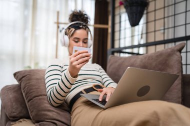 İnternetten alışveriş yapan bir kadının kredi kartı ve dizüstü bilgisayarı var. Evde genç yetişkin bir kadının evdeki kanepe yatağında otururken internetten alışveriş yaptığını görüyorlar.