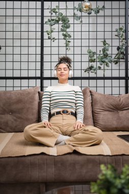Yetişkin bir beyaz kadın milenyum kulaklıklarını internet rehberli meditasyon için kullanıyor. Düşünce yogası yapıyor. Gözler kapalı bir şekilde kanepeye bakıyor.