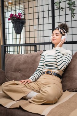 Yetişkin bir beyaz kadın milenyum kulaklıklarını internet rehberli meditasyon için kullanıyor. Düşünce yogası yapıyor. Gözler kapalı bir şekilde kanepeye bakıyor.