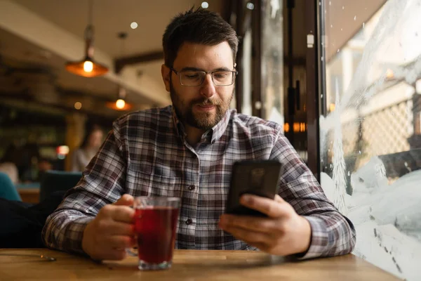 Adult caucasian man sitting alone at cafe use mobile smart phone for sms text messages or browse internet front view of one serious male with beard eyeglasses and cup of tea real people copy space