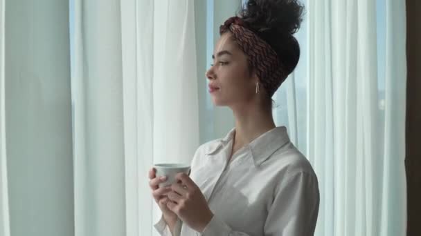 一人の女性幸せな白人女性が家の窓に立っているか 明るい日にホテルの部屋でコーヒーを飲みながら楽しむ毎日の朝のルーチン本物の人々は白いシャツを着ている — ストック動画