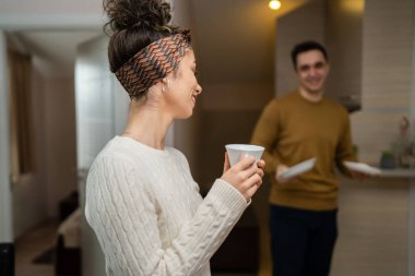 One woman young caucasian female standing in her apartment with cup of coffee while her husband or boyfriend is in the kitchen daily morning evening couple routine real people copy space