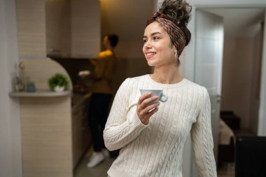 One woman young caucasian female standing in her apartment with cup of coffee while her husband or boyfriend is in the kitchen daily morning evening couple routine happy smile real people copy space
