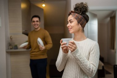 One woman young caucasian female standing in her apartment with cup of coffee while her husband or boyfriend is in the kitchen daily morning evening couple routine happy smile real people copy space