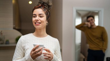 One woman young caucasian female standing in her apartment with cup of coffee while her husband or boyfriend stand in background daily morning evening couple routine real people copy space happy smile