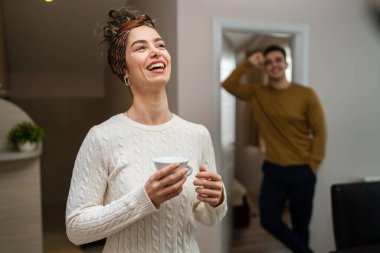 One woman young caucasian female standing in her apartment with cup of coffee while her husband or boyfriend stand in background daily morning evening couple routine real people copy space happy smile