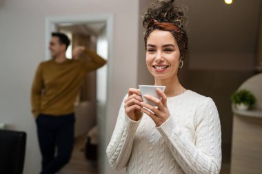 One woman young caucasian female standing in her apartment with cup of coffee while her husband or boyfriend stand in background daily morning evening couple routine real people copy space happy smile