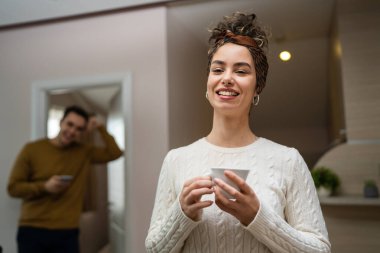 One woman young caucasian female standing in her apartment with cup of coffee while her husband or boyfriend stand in background daily morning evening couple routine real people copy space happy smile