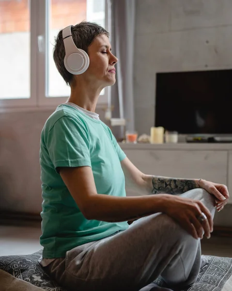 one woman senior caucasian female using headphones for online guided meditation practicing mindfulness yoga with eyes closed on the floor at home real people self care concept copy space