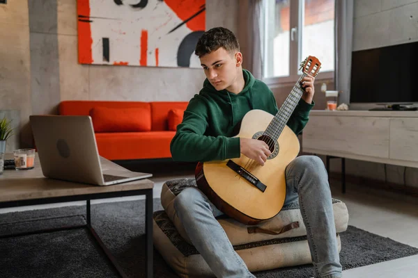 One young man Caucasian teenager sit at home in room playing guitar use laptop computer to learn for online lesson or course tutorial copy space