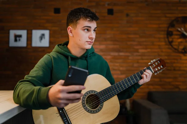 One young man Caucasian teenager sit at home in room playing guitar use smartphone mobile phone to learn for online lesson or course tutorial copy space