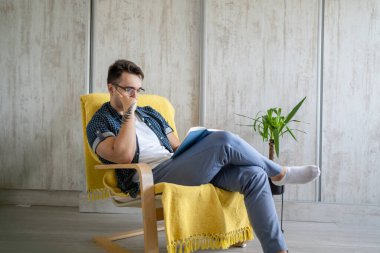 Kafkasyalı genç bir erkek, evde bir sandalyede oturmuş kitap okuyor mavi gömlek giyiyor ve gözlük takıyor. Düşünme, boş zaman kavramı, konsept eğitim alanı fotokopisi düşünüyor.