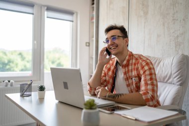 Beyaz tenli bir erkek çalışma masasındaki ofiste oturmuş cep telefonuyla sohbet ederken sohbet ediyor. Gömlek ve gözlük takıyor. Mutlu bir gülümseme takınıyor.