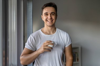 One man young adult male caucasian with brown hair standing by the window at home in morning day confident looking to the camera happy smile confident wear white shirt with cup of coffee copy space