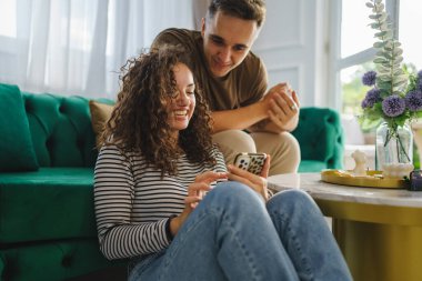 caucasian woman using mobile phone for online sms chat or internet browsing while her boyfriend or husband adult male sit beside at home in bright room real people couple leisure concept
