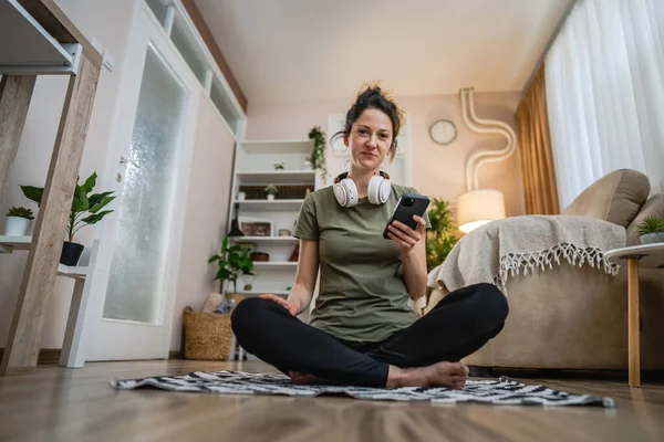1人の女性はヘッドフォンで自宅の床に座っている音楽を聴いたり インターネットコピースペース上の彼女のスマートフォンの携帯電話上のポッドキャストオンライン瞑想やオーディオブックをオンラインで検索するためにスマートフォンを使用しています — ストック写真