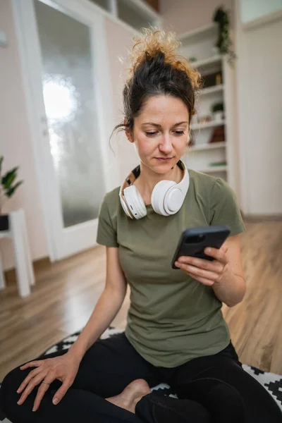 1人の女性はヘッドフォンで自宅の床に座っている音楽を聴いたり インターネットコピースペース上の彼女のスマートフォンの携帯電話上のポッドキャストオンライン瞑想やオーディオブックをオンラインで検索するためにスマートフォンを使用しています — ストック写真